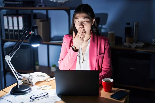 Chinese young woman working at the office at night bored yawning tired covering mouth with hand. restless and sleepiness.