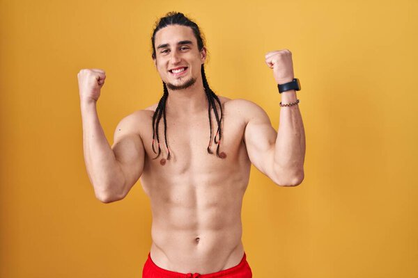 Латиноамериканец с длинными волосами, стоящий без рубашки на желтом фоне, радовался и удивлялся успеху с поднятыми руками и закрытыми глазами. победитель концепция. 