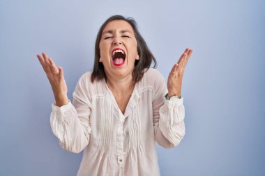 Orta yaşlı İspanyol kadın mavi arka planda ayakta duruyor ve başarı için çıldırıyor kollarını kaldırmış ve gözlerini kapatmış heyecanla haykırıyor. Kazanan konsept 