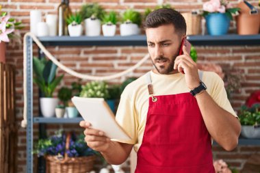 Young hispanic man florist talking on smartphone reading book at florist