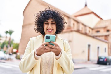 Orta Doğulu genç kadın güler yüzlü, kendine güler yüzlü, akıllı telefon kullanıyor.