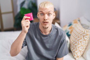 Kafkasyalı genç bir adam elinde prezervatifle yatakta korkmuş ve ağzı açık bir şekilde şaşırmış bir şekilde oturuyor. 