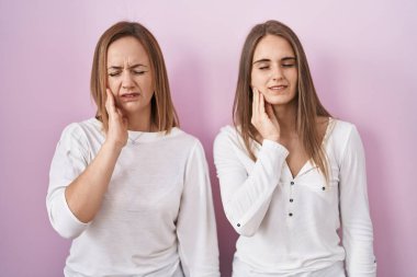 Orta yaşlı anne ve küçük kızı pembe arka planda durup diş ağrısı ya da diş hastalığından dolayı ağızlarına acı dolu bir ifadeyle dokunuyorlar. dişçi 