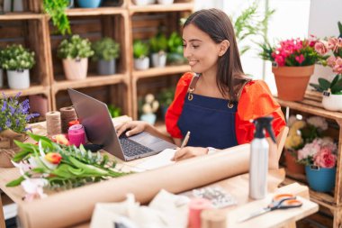 Young beautiful hispanic woman florist using laptop writing on notebook at florist