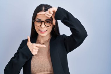 Young brunette woman standing over blue background smiling making frame with hands and fingers with happy face. creativity and photography concept. 