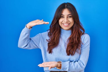 Mavi arka planda duran Latin kökenli genç bir kadın. Ellerinde büyük ve büyük bir işaret, ölçü sembolü var. Kameraya bakıp gülümsüyor. ölçüm kavramı. 