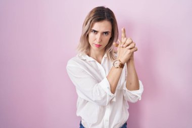 Pembe arka planda duran genç ve güzel bir kadın sembolik bir silahla el hareketleriyle cinayet silahlarını ateşleme oyunu oynuyor, kızgın bir yüz. 