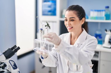 Young beautiful hispanic woman scientist holding test tubes with flowers at laboratory