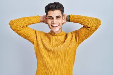 Young hispanic man standing over blue background relaxing and stretching, arms and hands behind head and neck smiling happy 