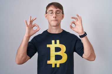 Bitcoin tişört giyen beyaz sarışın adam rahatlamış ve parmaklarıyla meditasyon hareketi yaparken gözleri kapalı gülümsüyor. yoga konsepti. 