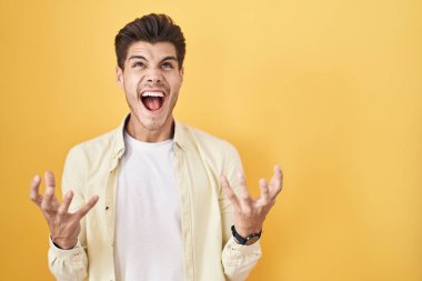 Young hispanic man standing over yellow background crazy and mad shouting and yelling with aggressive expression and arms raised. frustration concept. 