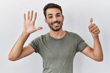 Beyaz arka plan üzerine gündelik tişört giyen, sakallı genç bir İspanyol. Altıncı parmaklarıyla işaret ederken kendinden emin ve mutlu gülümsüyor.. 