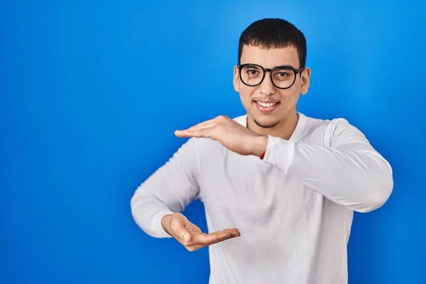 年轻的阿拉伯男子 身穿宽松的白色衬衫 戴着眼镜 手举着大号和大号的标志 表示尺寸的符号 微笑着看着摄像机 计量概念 — 图库照片