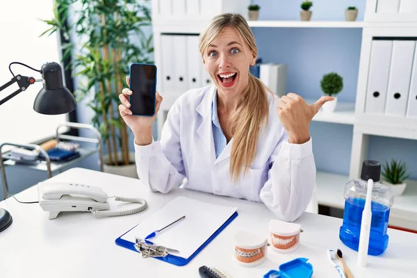 Beautiful Dentist Woman Showing Smartphone Screen Pointing Thumb Side Smiling — Stock Photo, Image