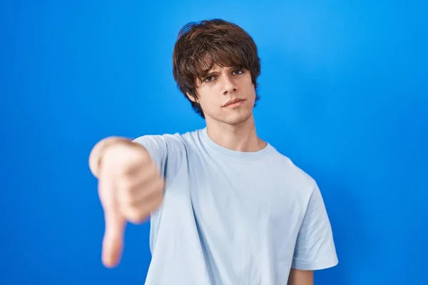 西班牙裔年轻人站在蓝色的背景上 满脸愁容 怒气冲冲 以大拇指垂下的姿势表示拒绝和消极 不好的表达方式 — 图库照片