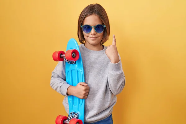小さなヒスパニック系の男の子を着てサングラスを保持スケート驚きでアイデアや質問を指差す指で幸せな顔 ナンバーワン — ストック写真