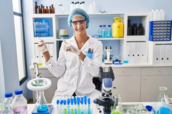 Brunette woman working at scientist laboratory smiling and looking at the camera pointing with two hands and fingers to the side.