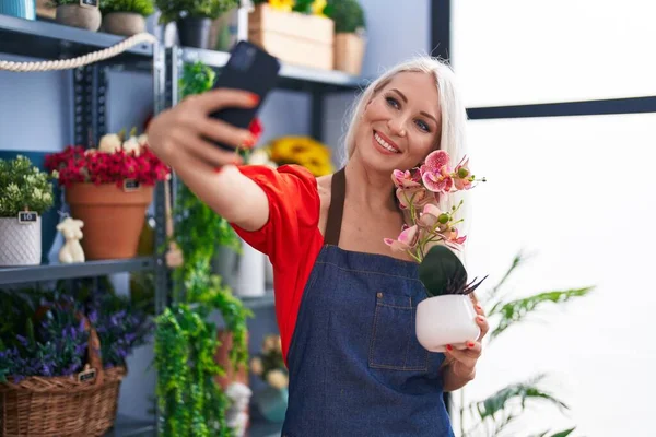 年轻的金发碧眼的女花匠在花店的智能手机手持设备旁自拍 — 图库照片