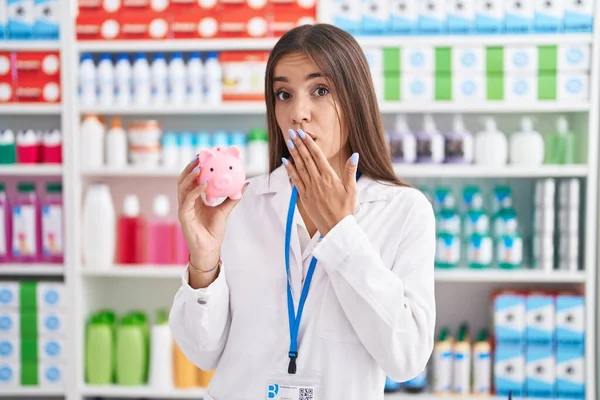 薬局で働いている若いブルネットの女性は 手で口を覆い ショックを受け 間違いを恐れて貯金箱を持っています 驚きの表情 — ストック写真