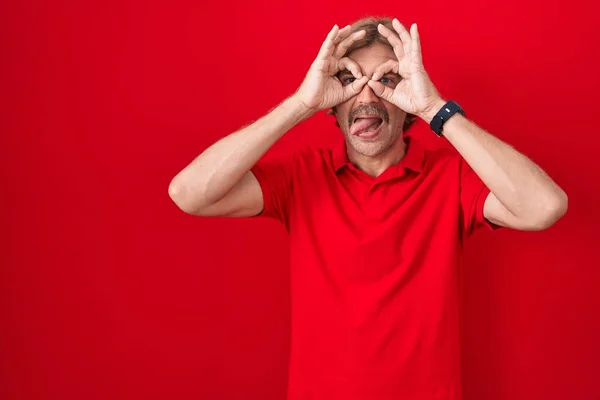 Caucasian man with mustache standing over red background doing ok gesture like binoculars sticking tongue out, eyes looking through fingers. crazy expression.