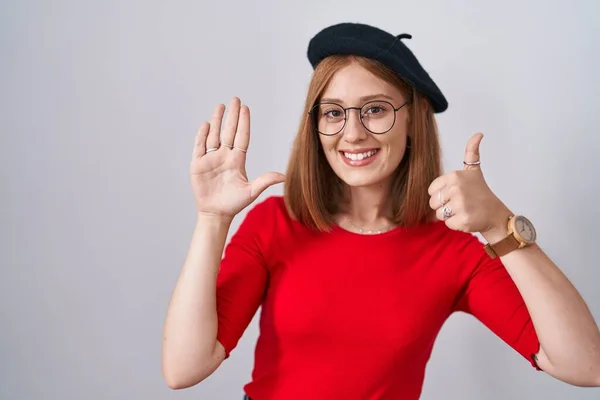 年轻的红头发女人站在那里 戴着眼镜 头戴贝雷帽 手握六号手指 自信而快乐地微笑着 — 图库照片