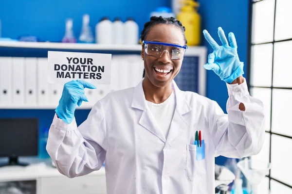Beautiful black woman working at scientist laboratory holding your donation matters banner doing ok sign with fingers, smiling friendly gesturing excellent symbol