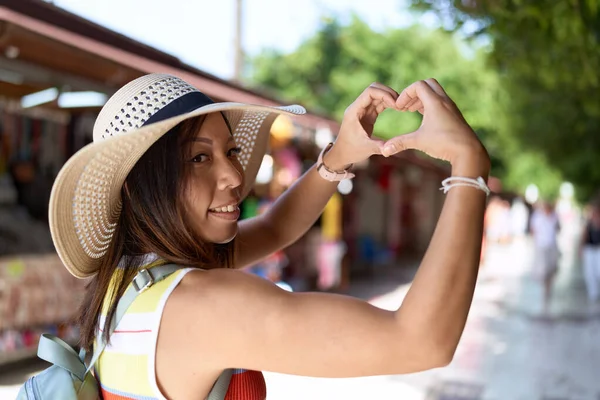 Young asian woman tourist smiling confident doing heart gesture with hands at street market