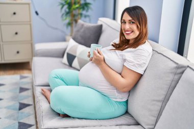 Young pregnant woman drinking coffee sitting on sofa at home