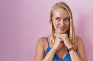 Young caucasian woman standing over pink background laughing nervous and excited with hands on chin looking to the side 