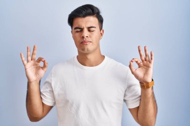Mavi arka planda duran İspanyol adam rahatlamış ve gözleri kapalı gülümsüyor parmaklarıyla meditasyon hareketi yapıyor. yoga konsepti. 