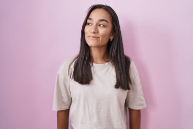 Young hispanic woman standing over pink background smiling looking to the side and staring away thinking. 