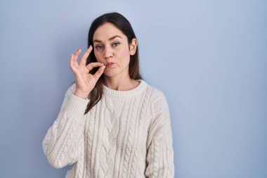 Young brunette woman standing over blue background mouth and lips shut as zip with fingers. secret and silent, taboo talking 