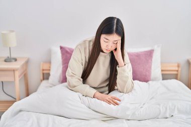 Çinli kadın yatak odasında ciddi bir ifadeyle yatakta oturuyor.