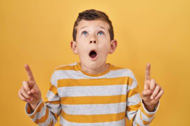 Sarı arka plan üzerinde duran genç beyaz çocuk şaşırmış ve şaşırmış bir şekilde yukarı baktı ve parmaklarıyla işaret etti ve kollarını kaldırdı.. 