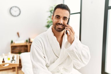 Young hispanic man wearing bathrobe talking on the smartphone at beauty center
