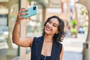 Genç ve güzel İspanyol kadın gülümsüyor. Kendine güveni tam. Sokakta akıllı telefonun yanında selfie çekiyor.