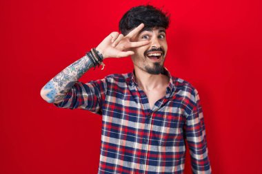 Kırmızı arka planda sakallı genç İspanyol adam barış sembolü yapıyor. Yüzünde parmakları, gülümseyerek zaferi gösteriyor. 