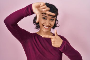 Pembe arka planda duran güzel bir kadın gülümsüyor ve mutlu yüzlü eller ve parmaklarla çerçeve yapıyor. yaratıcılık ve fotoğrafçılık kavramı. 