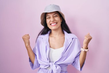 Pembe arka planda duran genç İspanyol kadın şapkasını takıyor. Şaşırmış ve şaşırmış bir şekilde kolları havada ve gözleri kapalı bir şekilde başarıyı kutluyor. Kazanan konsept. 