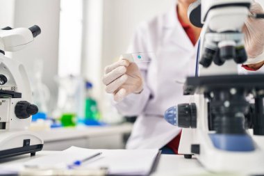 Laboratuvarda mikroskop kullanan orta yaşlı İspanyol kadın bilim adamı üniforması giyiyor.