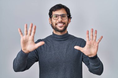 Yakışıklı Latin adam izole edilmiş arka planda duruyor ve 10 numarayı gösterip işaret ederken kendinden emin ve mutlu gülümsüyor.. 