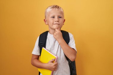 Little caucasian boy wearing student backpack and holding book serious face thinking about question with hand on chin, thoughtful about confusing idea 