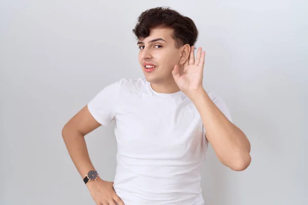 噂やゴシップに耳を傾ける耳の上に手で笑みを浮かべてカジュアルな白いTシャツを着て若い非バイナリ男 聴覚障害の概念 — ストック写真