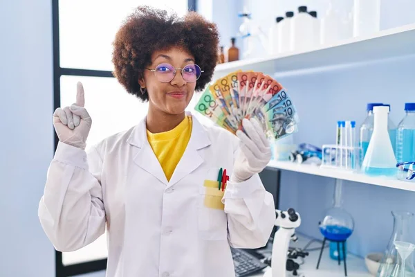 幸せな顔をしたアイデアや質問を指差す指で笑顔でお金を持っている科学研究所で働いている若いアフリカ系アメリカ人女性 ナンバーワン — ストック写真