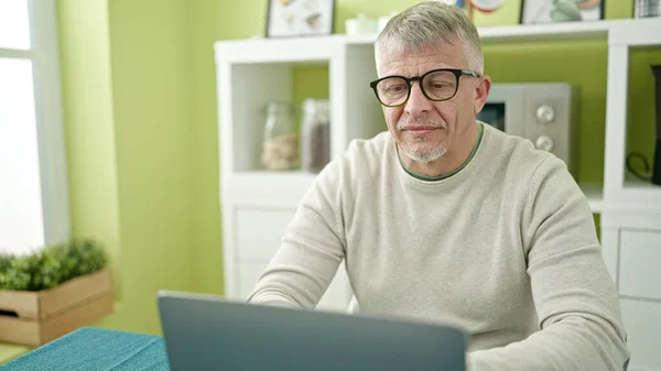 中年白发男子坐在家里的桌子上使用笔记本电脑 — 图库照片