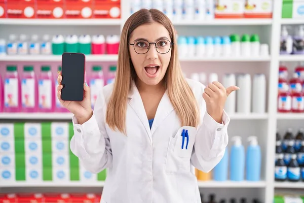 Young blonde woman working at pharmacy drugstore showing smartphone screen pointing thumb up to the side smiling happy with open mouth