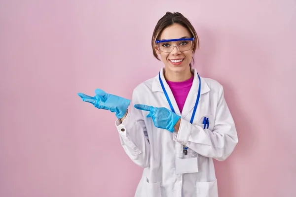 Hispanic woman wearing scientist uniform amazed and smiling to the camera while presenting with hand and pointing with finger.