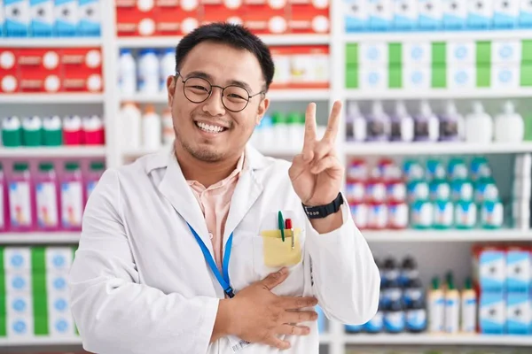 薬局で働く中国人青年が指で勝利サインをしてカメラに顔をウィンクして微笑む — ストック写真