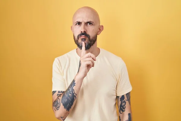 Hispanic man with tattoos standing over yellow background thinking concentrated about doubt with finger on chin and looking up wondering