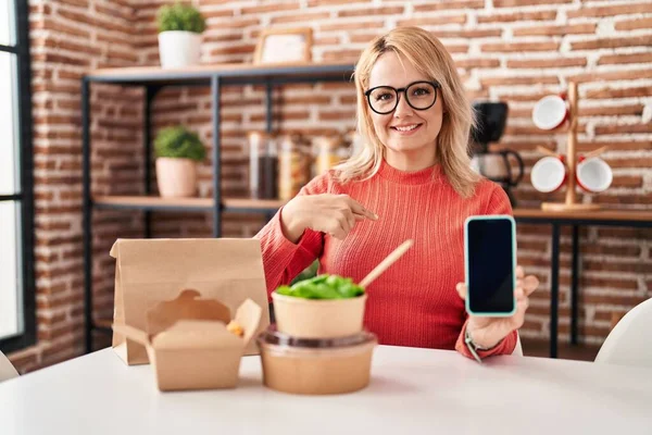 吃东西的金发女人把显示智能手机屏幕的食物拿走 指指点点着一个笑得开心而自豪的人 — 图库照片